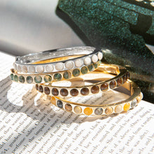 Load image into Gallery viewer, A stack of Semi Precious Stone Bangles rests on an open book, their vibrant inlays gently illuminated, casting delicate shadows across the pages. In the background, a glittery green surface adds to the scene&#39;s allure.
