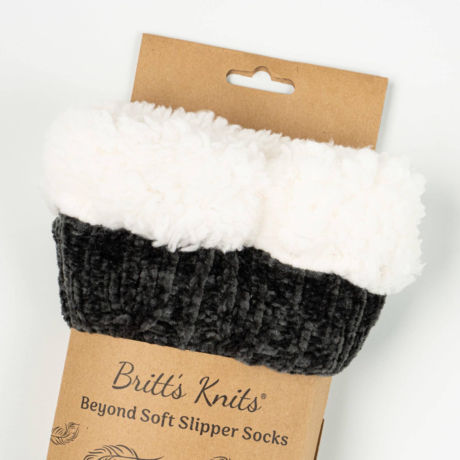 A pair of black Beyond Soft Slipper Socks, crafted with a cable knit design and lined with fluffy white material, is presented on a cardboard backing. The packaging proudly displays the label "Britt's Knits Beyond Soft Slipper Socks." These socks are designed with a ribbed texture and non-slip soles, offering both coziness and a soft appearance.