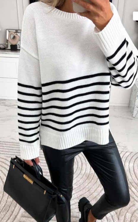 A person dressed in a Striped Sweater, characterized by its white fabric with black horizontal stripes and crafted in Italy, is holding a black handbag. They pair the sweater with black leather pants and shoes, surrounded by light-colored decor, while their phone is slightly visible in hand.