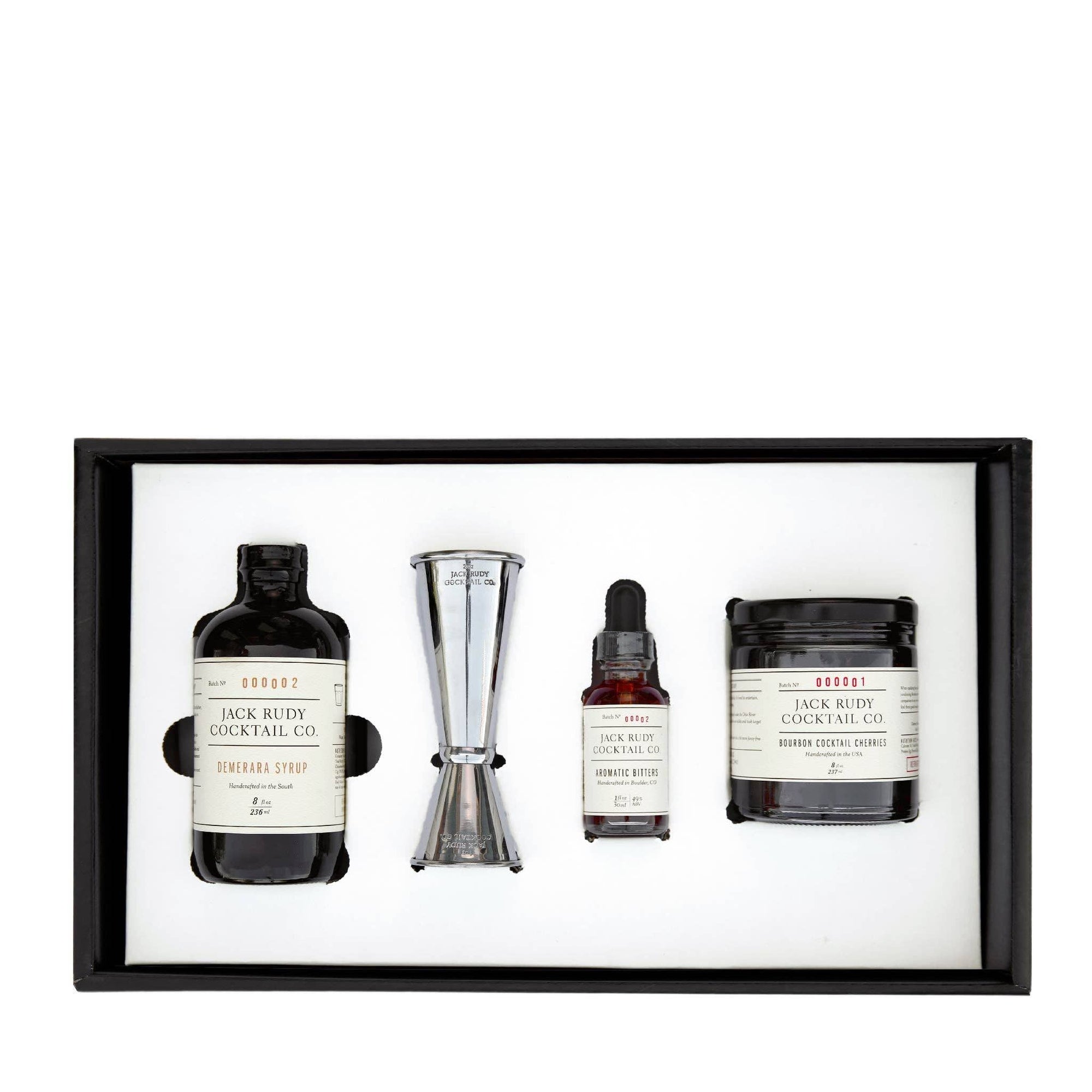 The Old Fashioned Kit - 4-pack, housed in a sleek black box, includes a bottle of syrup, a metal jigger, aromatic bitters, and cocktail cherries. Each item is labeled with the "Jack Rudy Cocktail Co." brand and features vintage-style packaging, all elegantly arranged on a pristine white background inside the box.