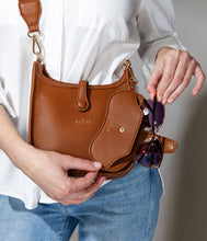 Load image into Gallery viewer, Wearing a white shirt and blue jeans, a person elegantly places sunglasses with purple lenses into an Eclipse Eyewear Case within a brown vegan leather handbag. The bag, featuring gold accents and logo, complements their light skin tone, blending style with practicality.
