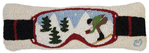 The Ski Goggles Pillow, a hand-hooked piece made from natural wool, showcases a rectangular design with ski goggles. Inside the goggles, there's a winter scene depicting a skier wearing brown pants and a green jacket navigating snowy slopes surrounded by pine trees and mountains. The black goggle strap is highlighted in red.