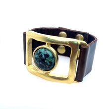 Load image into Gallery viewer, The &quot;Open Rectangle w Single Stone Leather Bracelet in Gold&quot; is a handcrafted piece from Brooklyn, featuring a dark brown leather strap with a large square frame in 18K gold, housing a round green and black speckled stone. It closes with two metal snaps on the strap.
