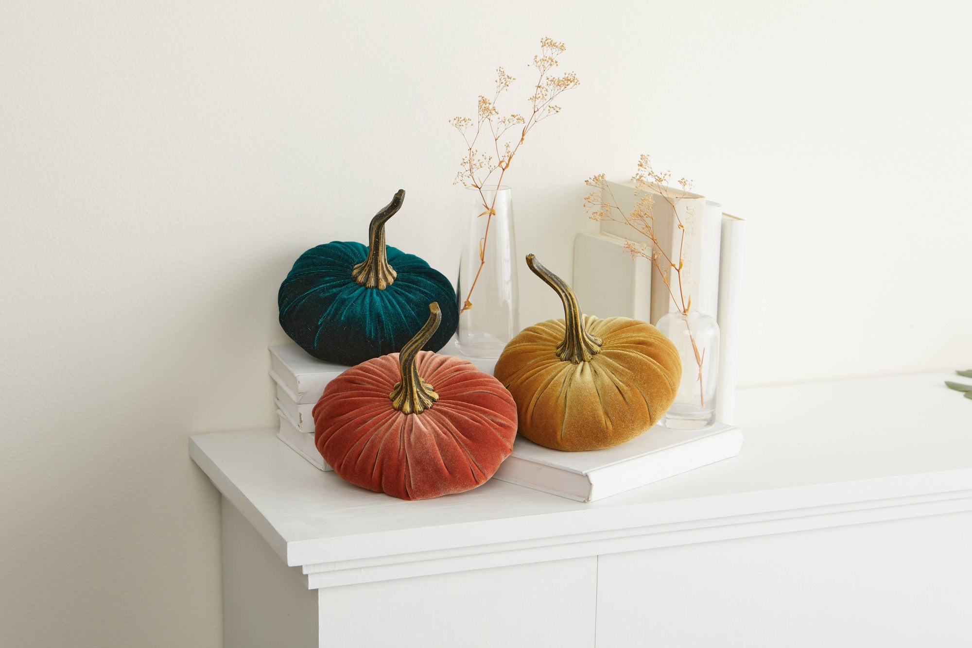 A trio of Handmade Large Velvet Pumpkins in the colors teal, orange, and mustard is elegantly displayed on white books that rest on top of a white cabinet. Each pumpkin features a realistic brown stem. In the background, there are two clear vases containing delicate dried flowers alongside several upright closed books, creating a minimalist fall décor ambiance.