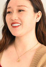 Load image into Gallery viewer, A woman with long brown hair is smiling and looking to the side, adorned with a 14K gold necklace featuring a circular pendant and Dignity Zircon Stud Earrings. Her red lipstick perfectly complements her tan sweater layered over a red top. The plain, light-colored background highlights her radiant appearance.
