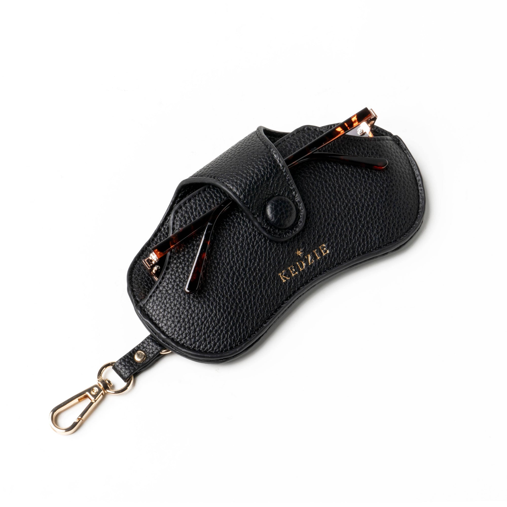 The Eclipse Eyewear Case is a black vegan leather case with a pebbled texture, featuring a flap closure, gold-toned snap button, and lobster clasp, with "KELZIE" embossed in gold. A peek of tortoiseshell glasses adds elegance to this sleek travel accessory.