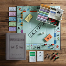 Load image into Gallery viewer, A Monopoly Vintage Bookshelf Edition set is artfully arranged on a wooden surface, featuring high-quality game pieces. The open board displays properties and spaces, with money, houses, hotels, dice, and cards meticulously organized alongside. A closed box labeled &quot;Monopoly Vintage Bookshelf Edition&quot; finishes the appealing setup for play.
