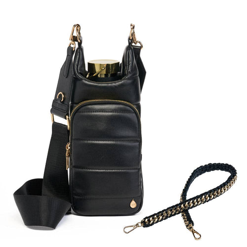 Introducing the Vegan Leather HydroBag® with Strap Bundle: a stylish black bag designed for hands-free wear and to hold your water bottle. It features a zippered front pocket and exquisite gold hardware, including a decorative charm at the bottom. Two detachable straps, one wide and plain, the other braided with gold and black detailing, complete its chic look.