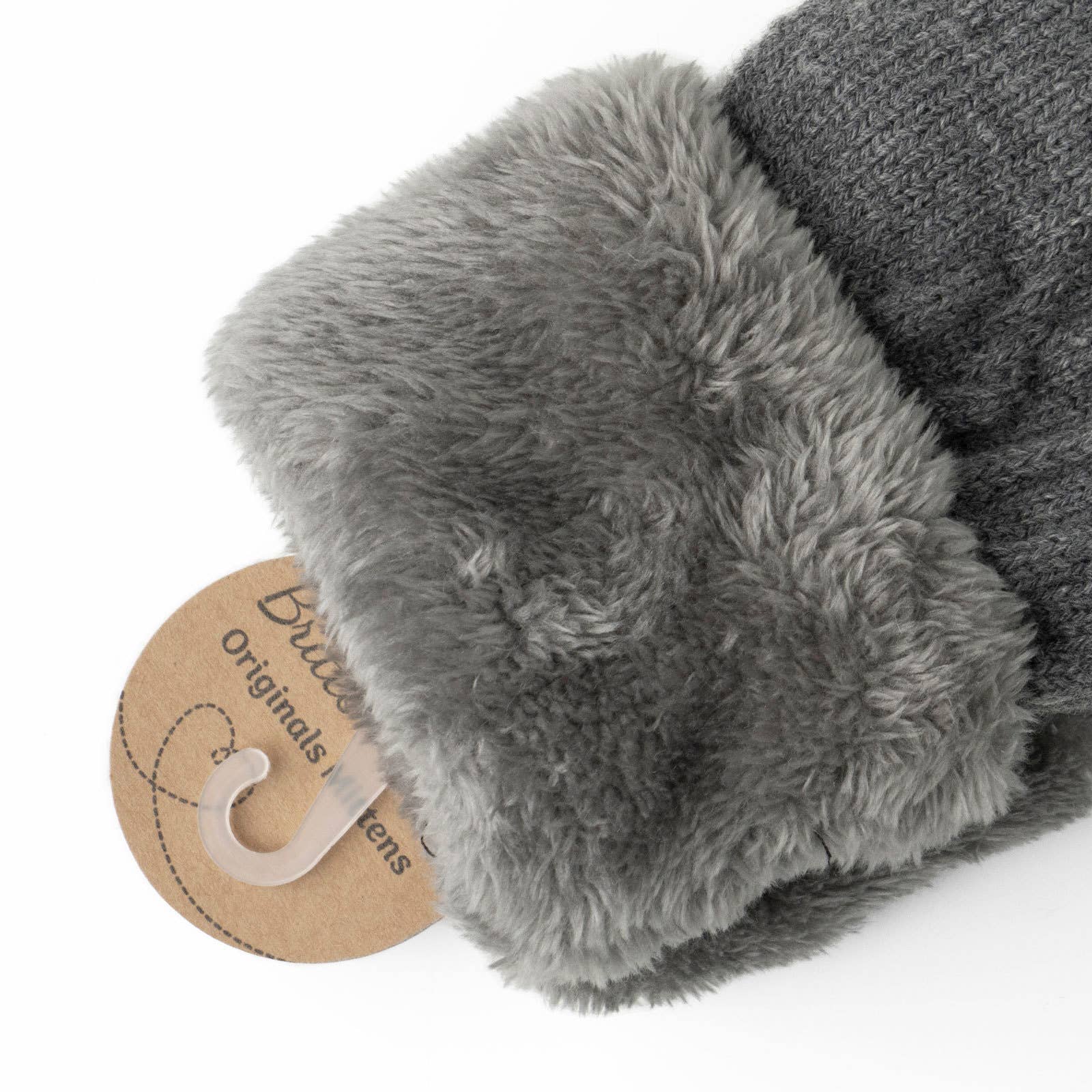 A close-up of the Cozy Cable Knit Mittens highlights their gray hue and features a snug cable knit design complemented by a soft, fluffy cuff. The mittens have an ultra-warm plush lining that looks incredibly inviting. A brown paper tag is partially visible, displaying text in a stylish font that underscores the comfort, warmth, and style perfect for cold weather.