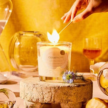 Load image into Gallery viewer, A hand lights a Champagne Candle 6 oz on a rustic stone surface, part of the Sparkling Collection. The background displays a carafe, amber liquid in a glass, and floral decor. Warm lighting creates a cozy atmosphere perfect for celebration.
