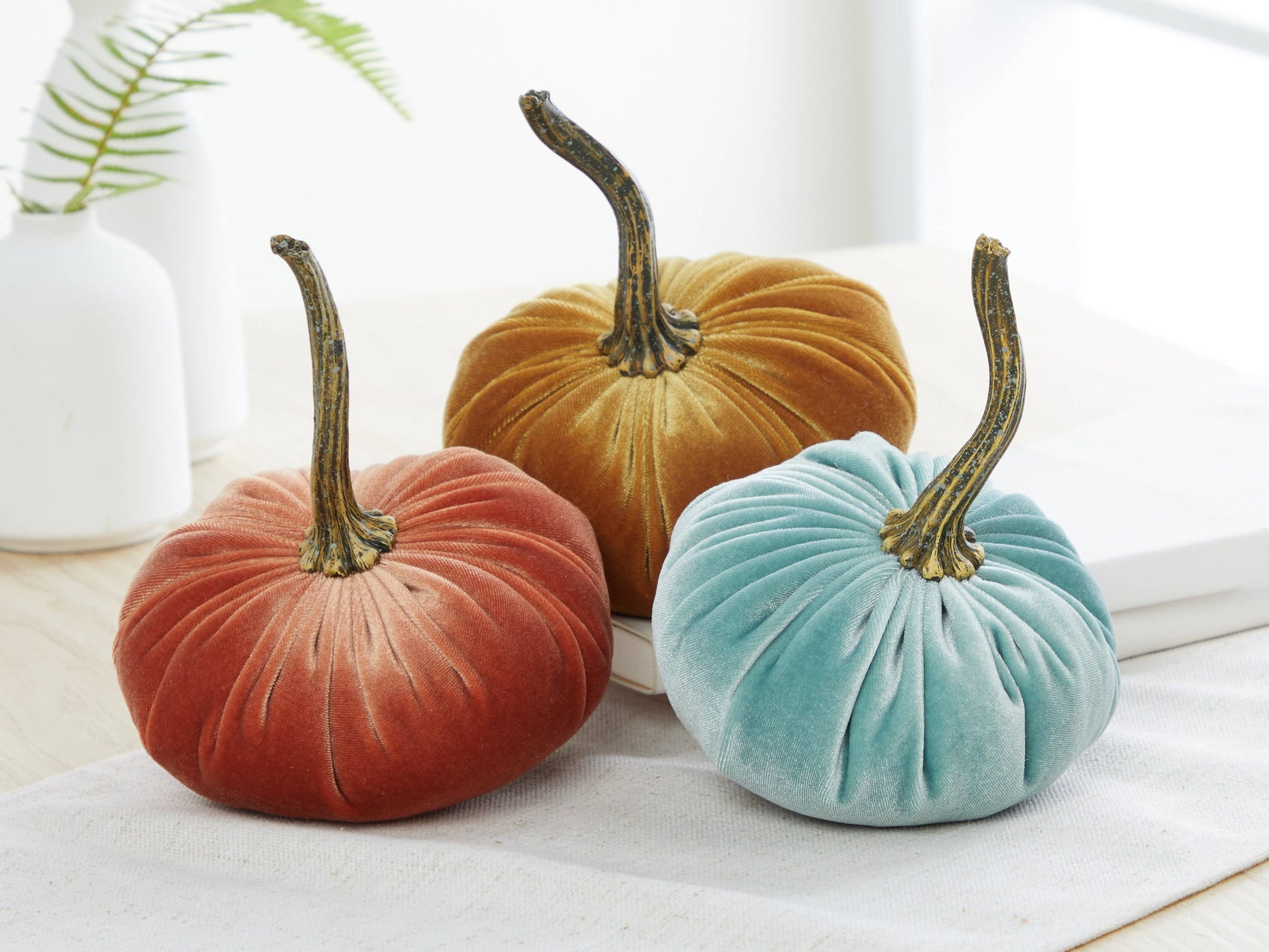 A trio of Handmade Small Velvet Pumpkins in rust, mustard, and teal are elegantly arranged on a wooden table. Each pumpkin boasts a dark brown stem. In the backdrop, a white vase filled with lush green fern leaves complements the display. The gentle lighting accentuates the warm, autumnal atmosphere, creating an ideal fall decor idea or wedding centerpiece.