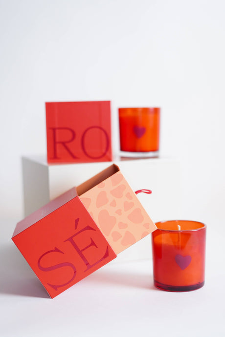 A Rosè V-Day Boxed Tumbler, featuring a red candle in a heart-patterned glass holder and emitting a subtle rosé fragrance, sits on white surface near a Valentine’s box with abstract hearts and 