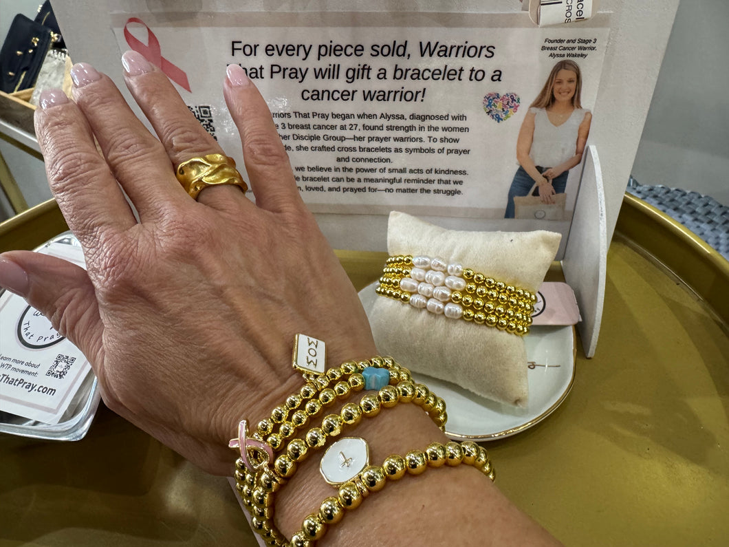 A Beaded Charm Bracelet featuring a pink ribbon charm graces a hand on a brass tabletop. A nearby sign encourages gifting these bracelets to cancer warriors, while two gold and pearl beaded bracelets rest on a pillow, and the hand is adorned with a gold ring and watch.