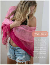 Load image into Gallery viewer, A woman wears a Wabi Silk Scarf in pink ombré over a black lace top and distressed denim shorts. The scarf is handspun and handwoven sustainable silk, dyed in a gemstone palette by artisans in Thailand, measuring 58 x 18 inches.
