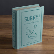 Load image into Gallery viewer, The image showcases the &quot;Sorry! Vintage Bookshelf Edition&quot; board game, elegantly packaged in a fabric-wrapped, book-like box. Adorned with the game&#39;s title and logo featuring two game pieces, it rests on a wooden surface. Suitable for ages 6 and up, it accommodates 2-4 players.
