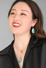 Load image into Gallery viewer, A woman wearing the It Is Well Necklace with a matching pendant beams as she gazes upward, donning 14K gold plated turquoise oval earrings. Her long hair cascades down, complemented by vivid red lipstick and winged eyeliner, set against a simple gray backdrop that exudes faith and peace.
