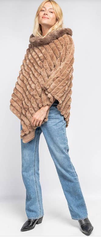 With long blonde hair, a person wears the stylish Faux Fur Poncho featuring a wide collar, offering both warmth and flair. They complement it with light blue jeans and black heeled boots. The outfit pops against the plain white background as they confidently lean to one side.