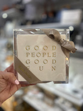 Load image into Gallery viewer, A hand holds the Cocktail Napkin Acrylic Tray Set - Year Round Entertaining, featuring decorative disposable napkins wrapped in a transparent box with a gold ribbon, making it an ideal hostess gift. The napkins are adorned with the words &quot;GOOD PEOPLE GOOD FUN&quot; printed in gold. In the blurred background, shelves display an array of other items.
