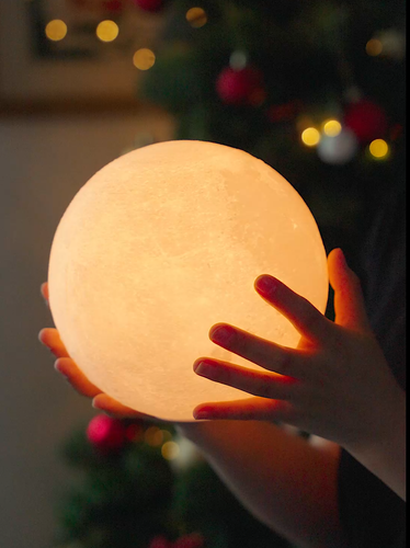 A person holds the Smart LunaSpin Lamp, a glowing spherical lamp that resembles a moon and features 3D-printed details, encircled by gentle hands. Blurred festive lights and decorations in the background suggest a holiday atmosphere, while the lamp's warm light creates a cozy and serene ambiance.