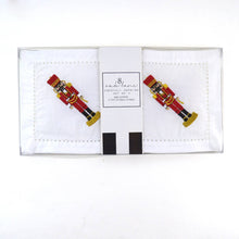 Load image into Gallery viewer, The Nutcracker Embroidered Cocktail Napkin Boxed Set includes a collection of white cocktail napkins made from 100% cotton, showcasing intricately embroidered red and gold nutcrackers. Packaged in a transparent box with the &quot;andSons&quot; label on top, each machine-washable napkin features a centrally placed nutcracker design with neat stitching.
