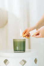 Load image into Gallery viewer, A person with neatly manicured nails lights the Juniper Berry &amp; Fir Soy Candle, resting on a white, glossy, round surface. Soft, natural light enhances the serene ambiance as smoke rises gently from the flame against a blurred, light background.
