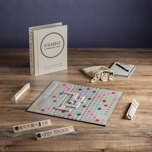 Load image into Gallery viewer, The image displays a Scrabble Vintage Bookshelf Edition game artfully arranged on a wooden table. The board showcases words created from high-quality game components, with racks featuring &quot;WORDS&quot; and &quot;VINTAGE.&quot; A beige box, score pad with pencil, and drawstring bag accompany the setup against a plain dark wall background.
