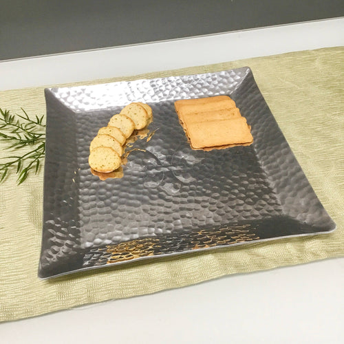 The beige cloth is elegantly adorned with the Hammered Aluminum Emblem Tray, featuring a silver finish. Resting atop the tray is a small stack of herb-infused round crackers, partially topped with mustard or sauce. Alongside them lies an orderly row of rectangular cookies, complemented by a sprig of rosemary for an added touch of elegance.