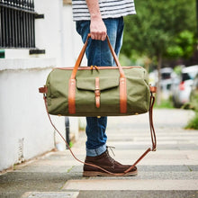 Load image into Gallery viewer, A person stands on a sidewalk holding a Leather Trim Weekend Travel Bag, featuring a spacious green design with elegant brown leather straps and details. They wear a striped shirt, blue jeans, and brown suede boots. The street is lined with trees, and parked cars are visible in the background.
