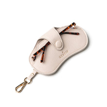 Load image into Gallery viewer, The Eclipse Eyewear Case is a chic travel accessory made of beige vegan leather with a textured finish and button clasp. It features two tortoiseshell-patterned temples peeking out, a gold-tone clip at the bottom left, and &quot;KEDZIE&quot; embossed in gold on the front.
