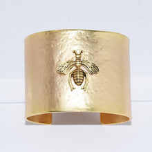 Load image into Gallery viewer, The Bee Design Hammered Cuff Bracelet features a broad, textured gold band with an intricate bee motif at its center. The bee&#39;s detailed wings and body create a subtle contrast against the matte, hammered gold backdrop. Elegantly displayed upright against a plain white setting, this bracelet exudes sophistication and grace.
