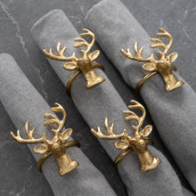 Load image into Gallery viewer, Gold Deer Napkin Rings from the set of 4 adorn rolled gray napkins, resting on a dark stone surface. These rings, crafted from sustainable materials with intricate antlers and detailed deer faces, add a rustic charm to the table setting.
