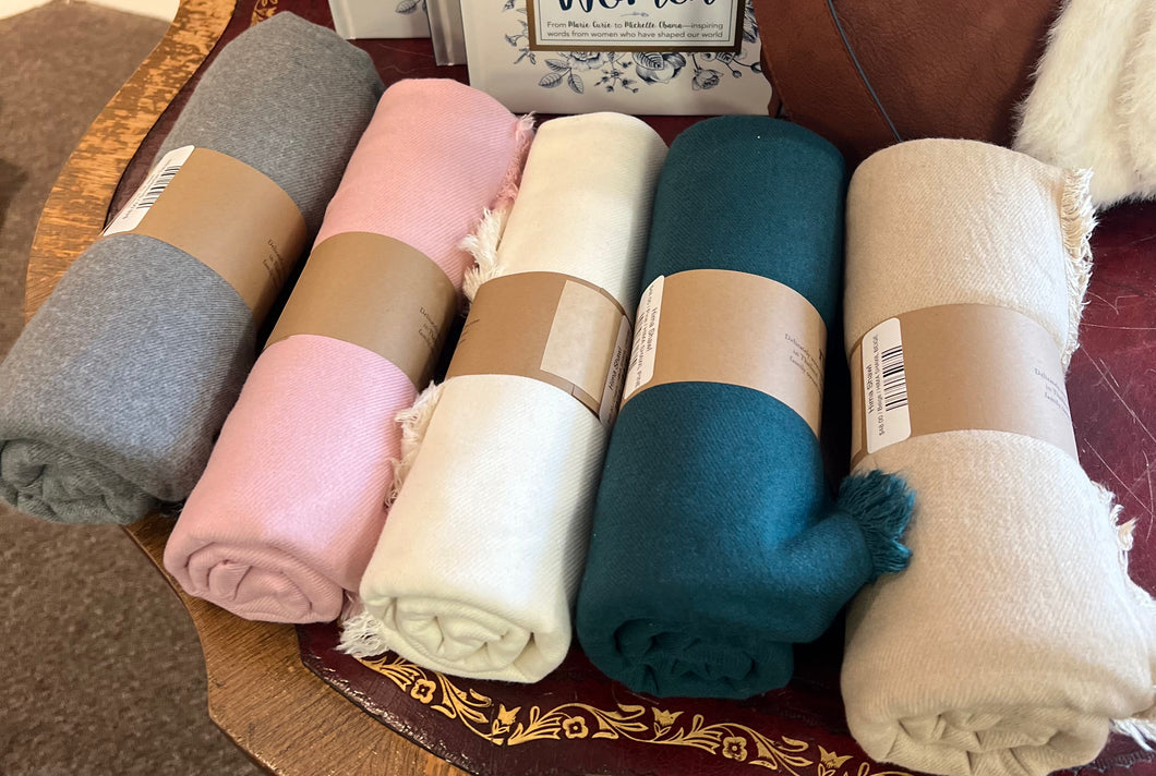 Five rolled fabric bundles in muted colors—gray, pink, white, teal, and beige—are neatly arranged on a decorative wooden table. Each Hima Large Scarf, Wrap, Shawl bundle is secured with a brown paper band featuring a label and barcode. The fabrics' fringed edges reflect their commitment to sustainable fashion.