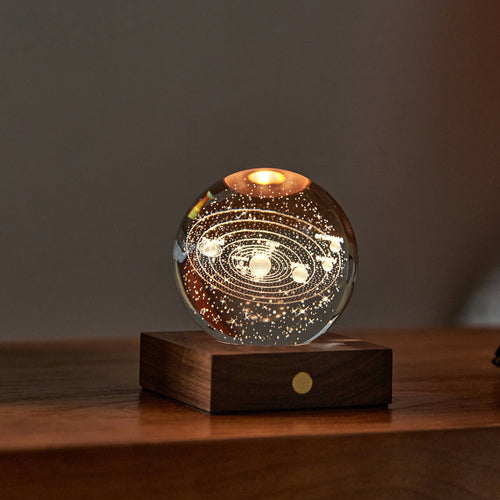 The Amber Crystal Light is a glass sphere night light featuring an illuminated solar system model with intricately etched planets and orbits inside. It sits on a wooden base, and the warm light accentuates its fine details, while the blurred dark background enhances this exquisite home décor piece's glow.