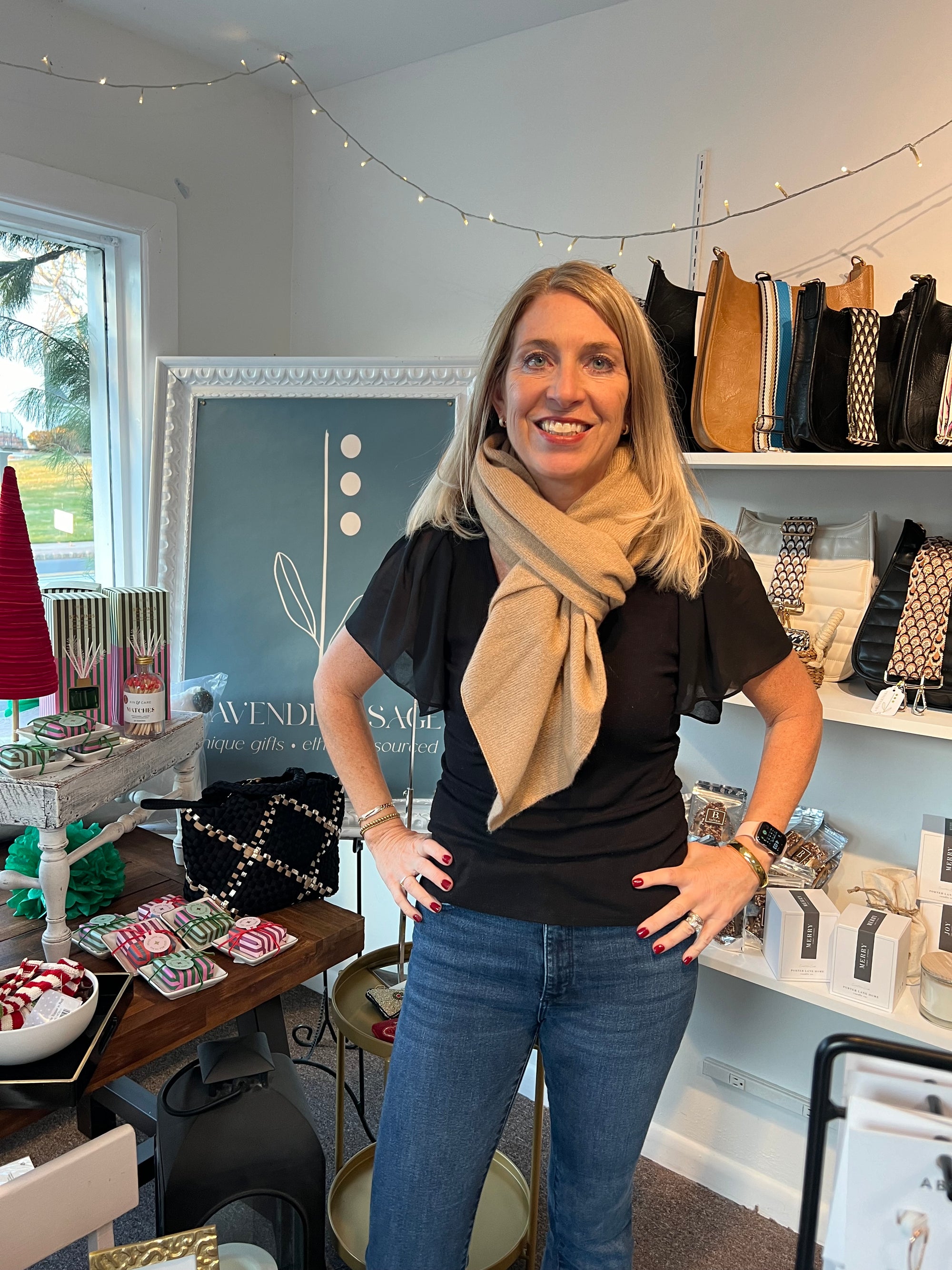 A woman with long blonde hair stands smiling in a shop, wearing a black top, blue jeans, and showcasing a tan scarf. Behind her are shelves displaying bags and accessories. A chalkboard sign highlights "One Size" items, including the Cashmere Poncho she models. String lights enhance the charm of the scene. 到着.