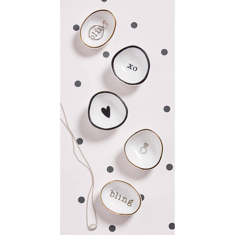 Four small white Ring Dishes with gold rims sit on a polka dot background. They feature black designs: "hi," "xo," a heart, and a ring with "bling." A silver necklace with a square pendant lies beside them, ideal for storing your treasures.