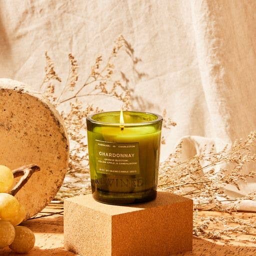 The Chardonnay Candle 6 oz from the Signature Collection sits on a cork block, its green wax gently flickering. Dried grasses and grapes surround it against a beige fabric backdrop, creating a cozy, rustic charm ideal for wine lovers.