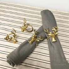 Load image into Gallery viewer, Gray napkins, made from sustainable materials, are elegantly rolled and adorned with Gold Deer Napkin Rings on a striped cloth of dark and light lines. Two additional handmade rings rest nearby, each featuring intricate antlers and a shiny finish from the set of four.
