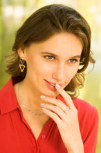 Load image into Gallery viewer, A woman with a contemplative look wears a red blouse and gold triangular earrings, complemented by the Ada Silver Heart Ring on her finger. Her shoulder-length brown hair frames her face as she thoughtfully places her fingers on her lips, against a softly blurred background of green and yellow hues.
