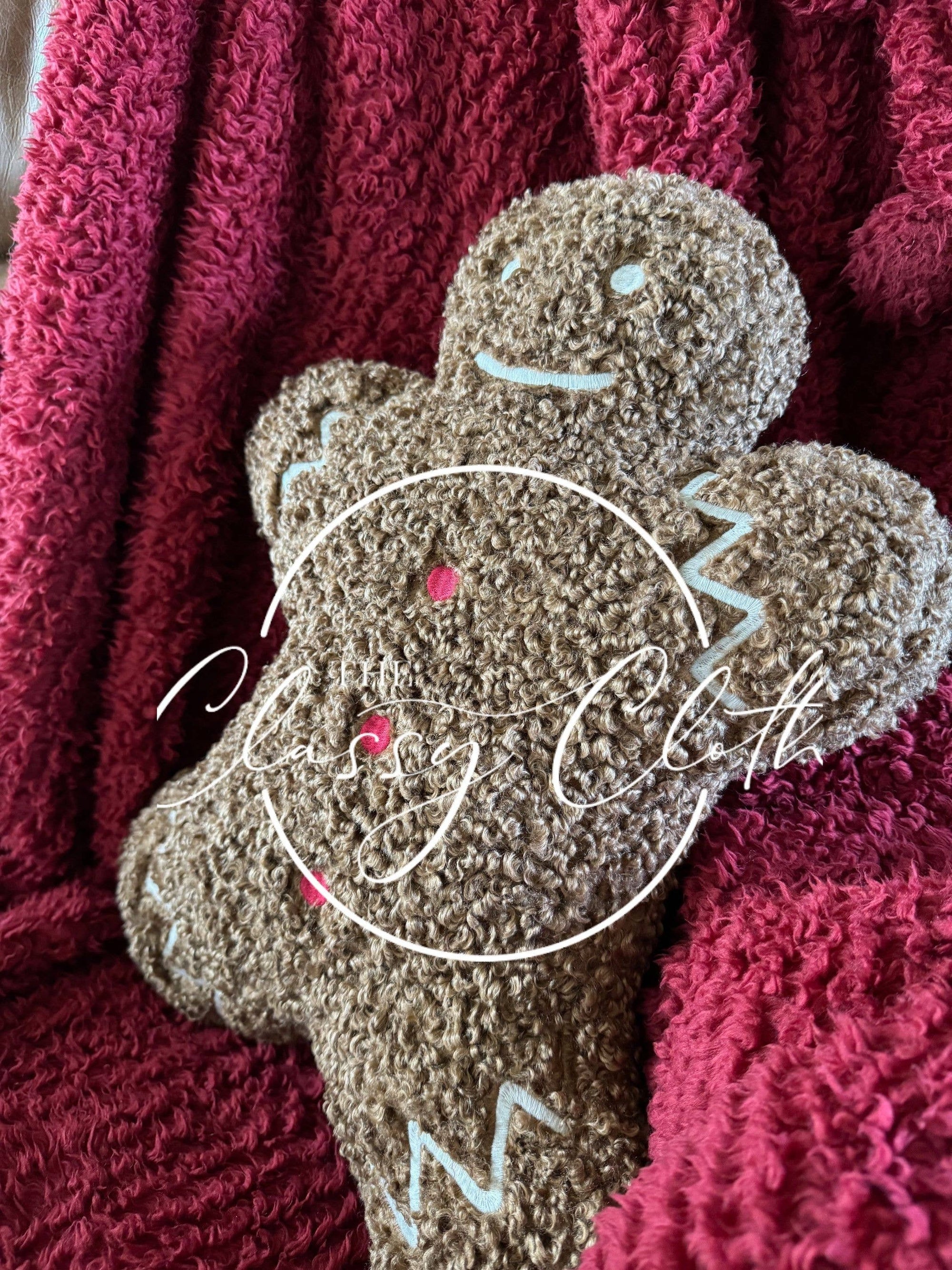 A textured Christmas Sherpa Pillow Gingerbread Man, featuring zigzag icing designs on its arms and legs, rests against a rich maroon, fluffy fabric backdrop. This Gingerbread Man design, perfect for holiday decorations, showcases a smiling face and pink spots for buttons. "Classy Cloth" is elegantly scripted over the image.