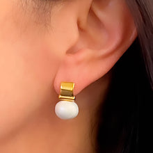 Load image into Gallery viewer, A close-up captures an ear wearing the Smooth + Classic Pebble Pearl Post Earrings, made from 14 karat gold and showcasing a white pearl. The rectangular gold section elegantly complements the pearl&#39;s dangle. Soft lighting accentuates its sheen, while hypoallergenic backing ensures comfort against hair and skin in the background.
