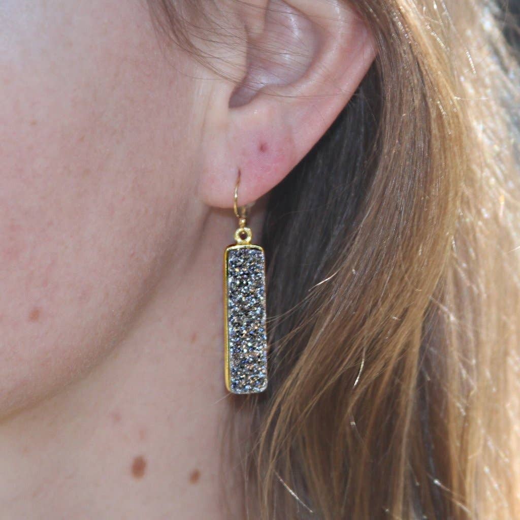 Close-up of a woman's ear adorned with a rectangular, gold-filled Druzy Baguette Earring that features a glittering silver bezel segment. The earring hangs from a delicate gold hook. Her light brown hair partially obscures the side of her face, while the background remains softly blurred.