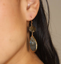 Load image into Gallery viewer, Close-up of a person wearing the Alexa Labradorite earrings, showcasing faceted pyrite above labradorite drops. The upper stone is spherical, and the lower is teardrop-shaped, both elegantly set in a gold frame with gold-filled ear wires. The person&#39;s dark hair and ear are visible against a neutral background.
