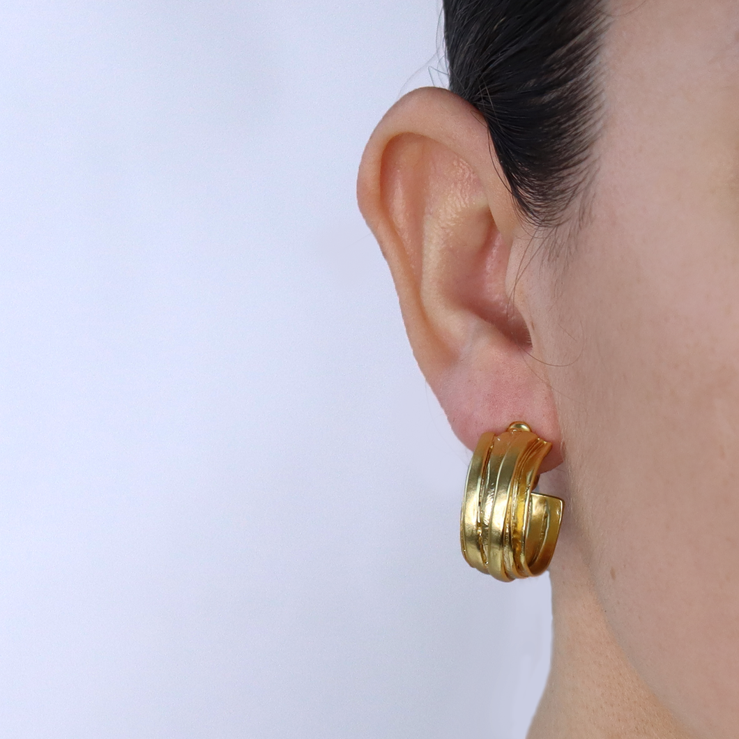 Close-up of a person's ear adorned with retro accessories, showcasing the Overlap Hoop Clip-On Earring. This gold piece consists of three intertwined bands, creating a layered look. Set against a plain white background that highlights the jewelry, only hints of the person's face and hair are visible, emphasizing their smooth skin.