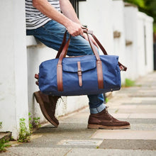 Load image into Gallery viewer, A person in blue jeans and brown boots leans against a white wall, holding a Leather Trim Weekend Travel Bag. The setting is an outdoor sidewalk with greenery in the background. The person wears a striped shirt, and their face is not visible.
