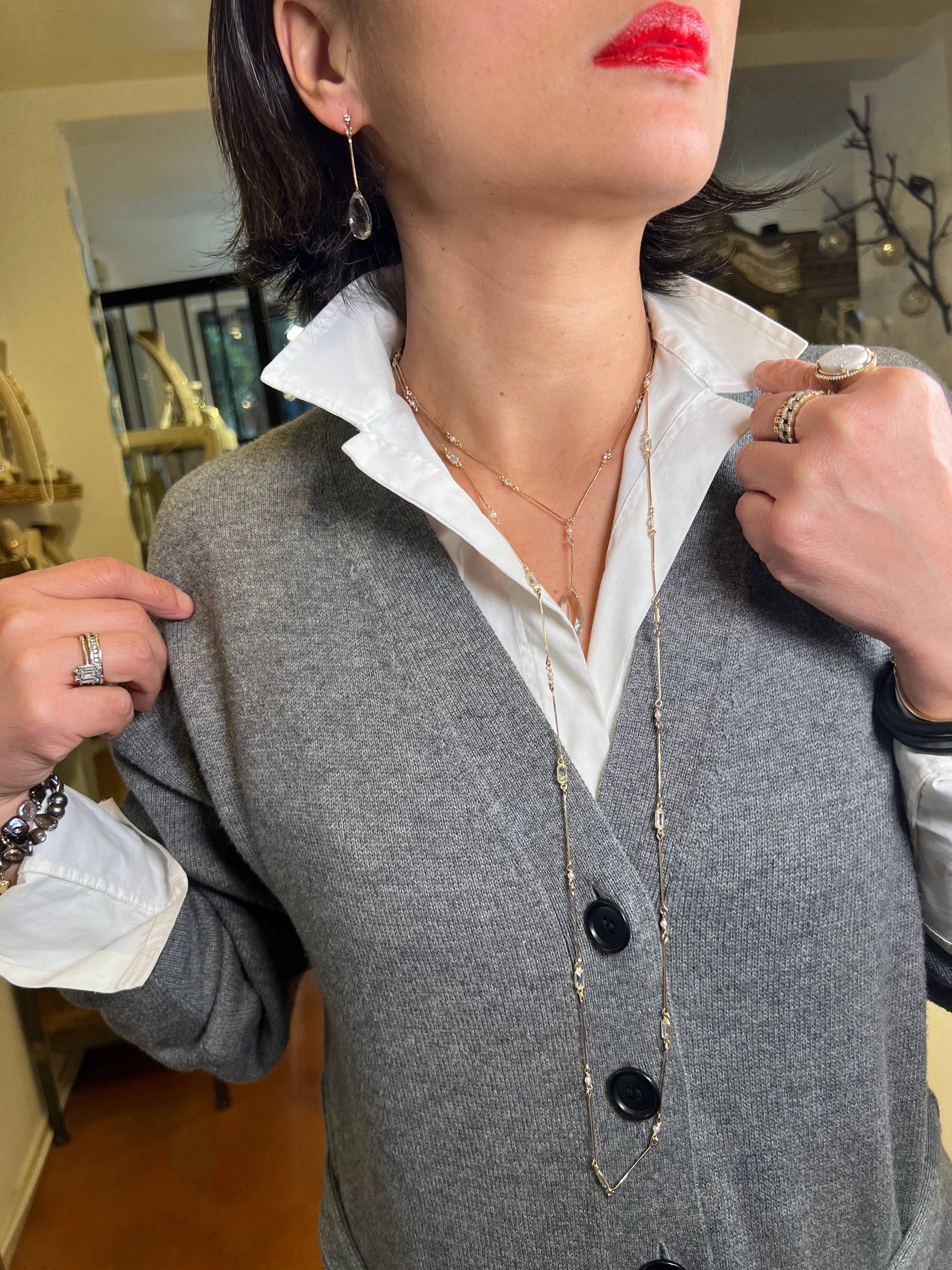 A person is wearing a white shirt with a popped collar under a gray sweater, accessorized with handcrafted layered gold earrings and the Green Amethyst Lariat Necklace. They have red lipstick and 14k gold-filled rings and bracelets. One hand adjusts the sweater while the other holds the collar against a backdrop of a staircase and decor.