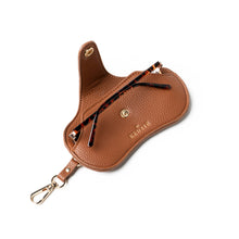 Load image into Gallery viewer, The Eclipse Eyewear Case is a brown textured vegan leather case with a gold snap button and &quot;KREWE&quot; embossed in gold. It includes a metal clasp for attachment, and showcases tortoiseshell-patterned glasses partially inside, ideal for stylish eyewear storage on the go.
