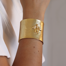 Load image into Gallery viewer, A close-up of a person&#39;s arm highlights the Bee Design Hammered Cuff Bracelet, featuring a wide, textured gold design with a raised bee motif. The hammered finish catches the light beautifully as the hand rests against a white fabric garment, set against a soft, blurred grey background.
