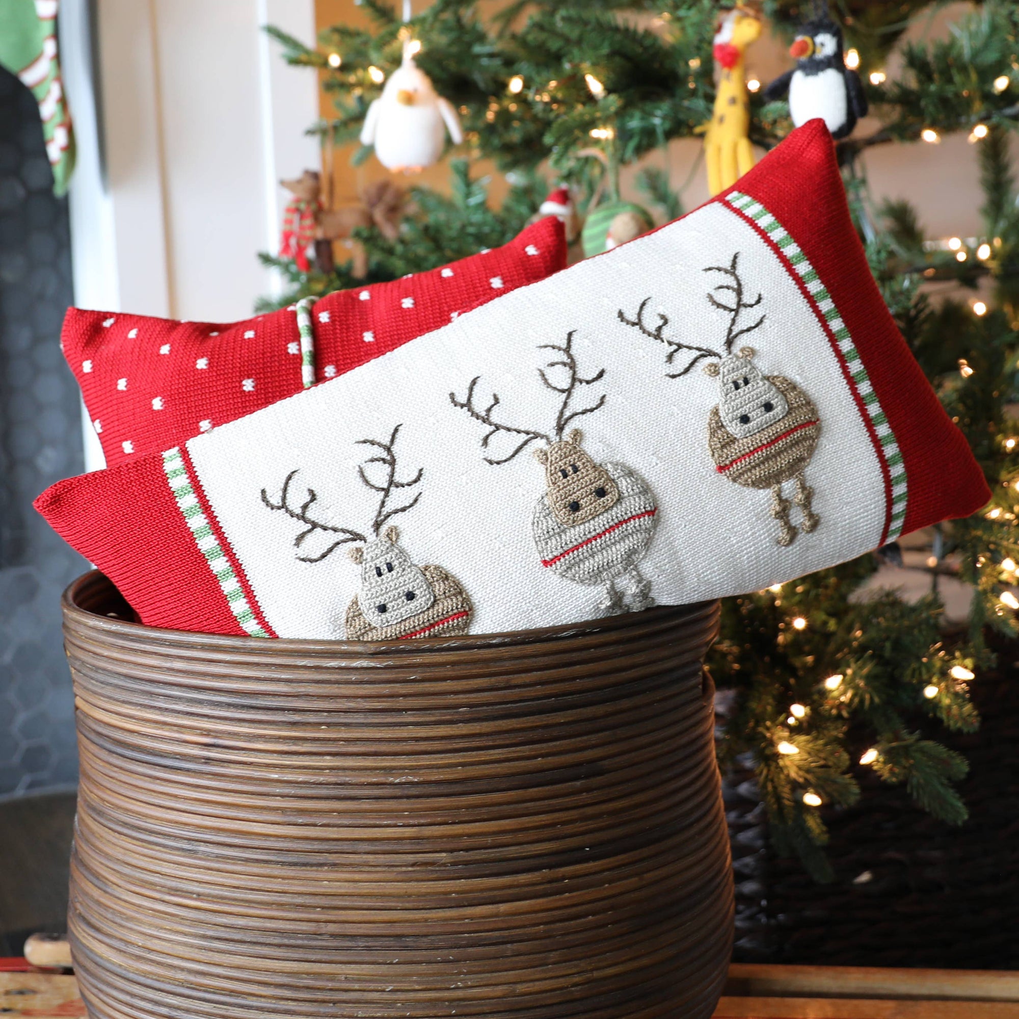A wicker basket contains two festive pillows, featuring the handmade Round Reindeer Lumbar Pillow. One pillow is red with white dots, while the other pillow features a design with three reindeer on a white background. Behind the basket, a decorated Christmas tree adorned with lights and ornaments adds to the warm and cozy indoor atmosphere.