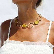 Load image into Gallery viewer, A woman in a white ruffled strapless top showcases the Vivid Gems Trio Necklace—a statement piece with large, oval, translucent yellow stones set in chunky gold. The bright white background accentuates the jewelry and her tanned skin.
