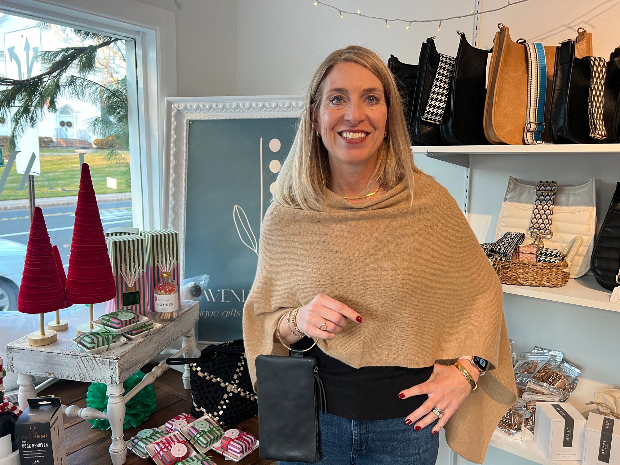 A woman with long blonde hair, dressed in a Cashmere Poncho and jeans, stands smiling in a store. She is holding a black clutch. Surrounding her are shelves showcasing bags in black and tan hues, while a table displays red cone trees, candles, and small gifts. Through the window is a view of greenery outside.